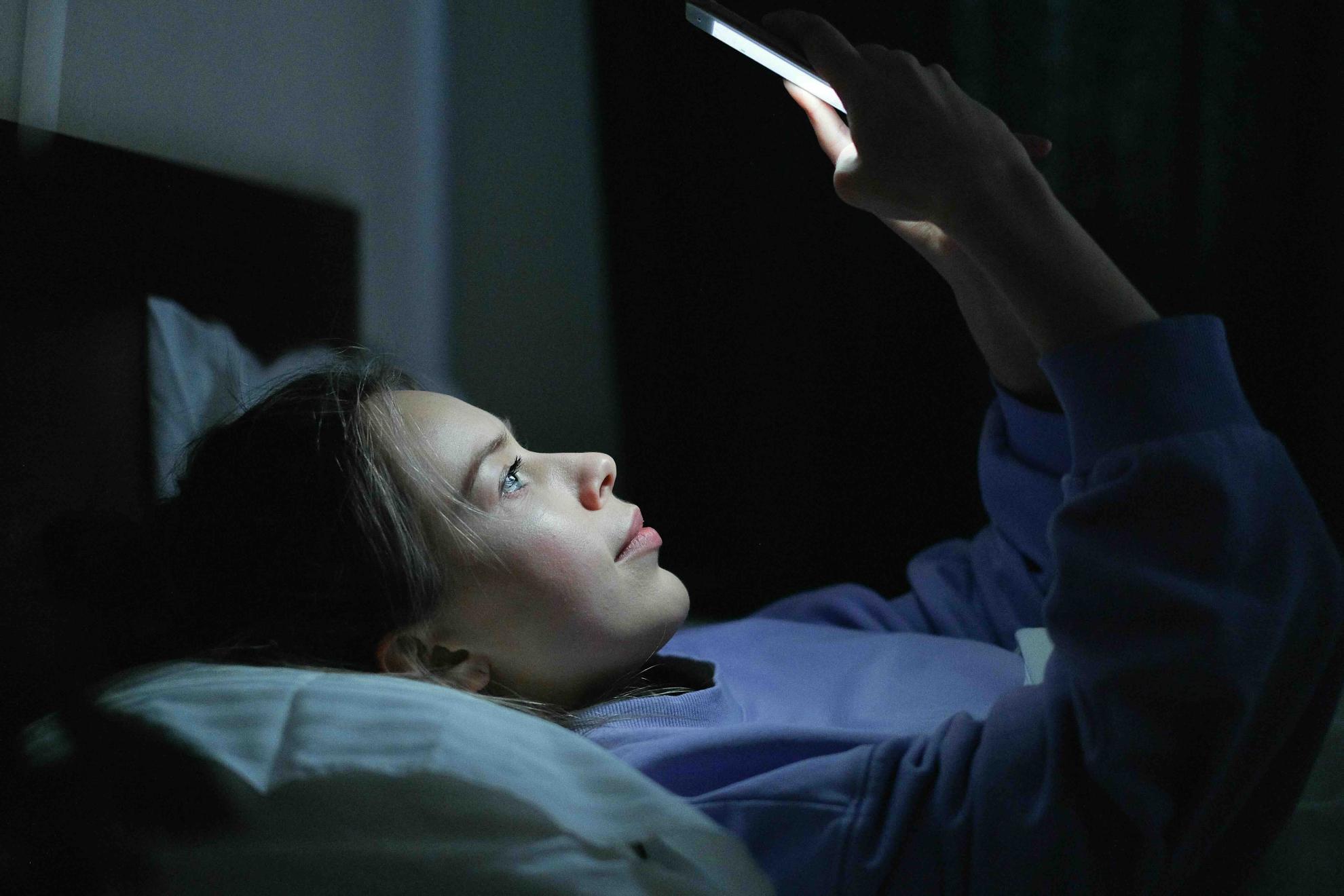 Una adolescente observa su teléfono móvil tumbada en la cama.